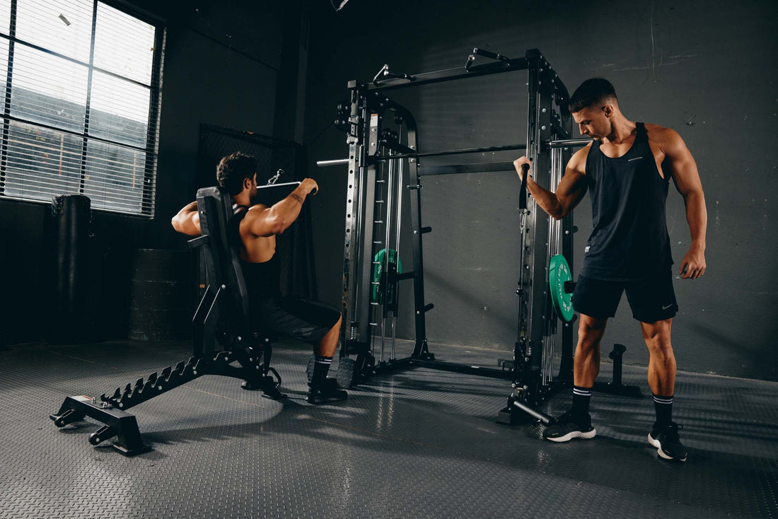 Man performing lat pulldowns on a bench while another uses a Smith machine.