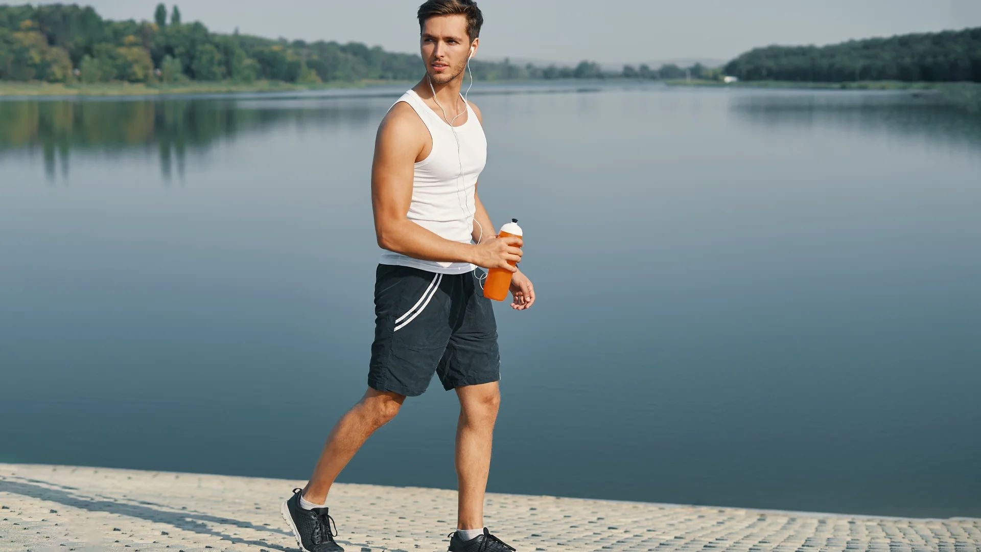 A young man on a fitness walk by a lake, carrying a water bottle and listening to music, promoting active lifestyle.