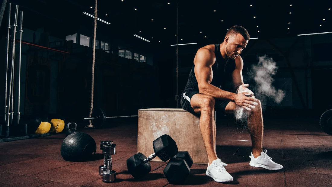 A man sitting on a box preparing for a workout in a gym, surrounded by dumbbells and fitness equipment, focusing on mental and physical preparation.v