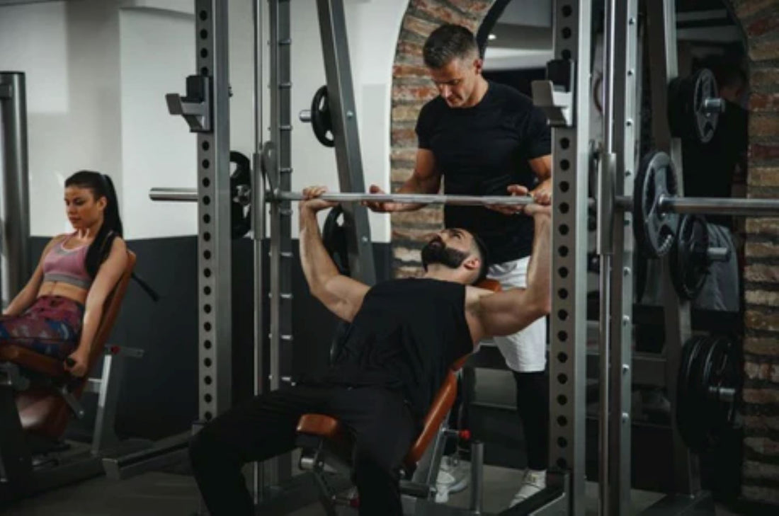 Man performing a bench press on a Smith machine with a trainer's assistance.