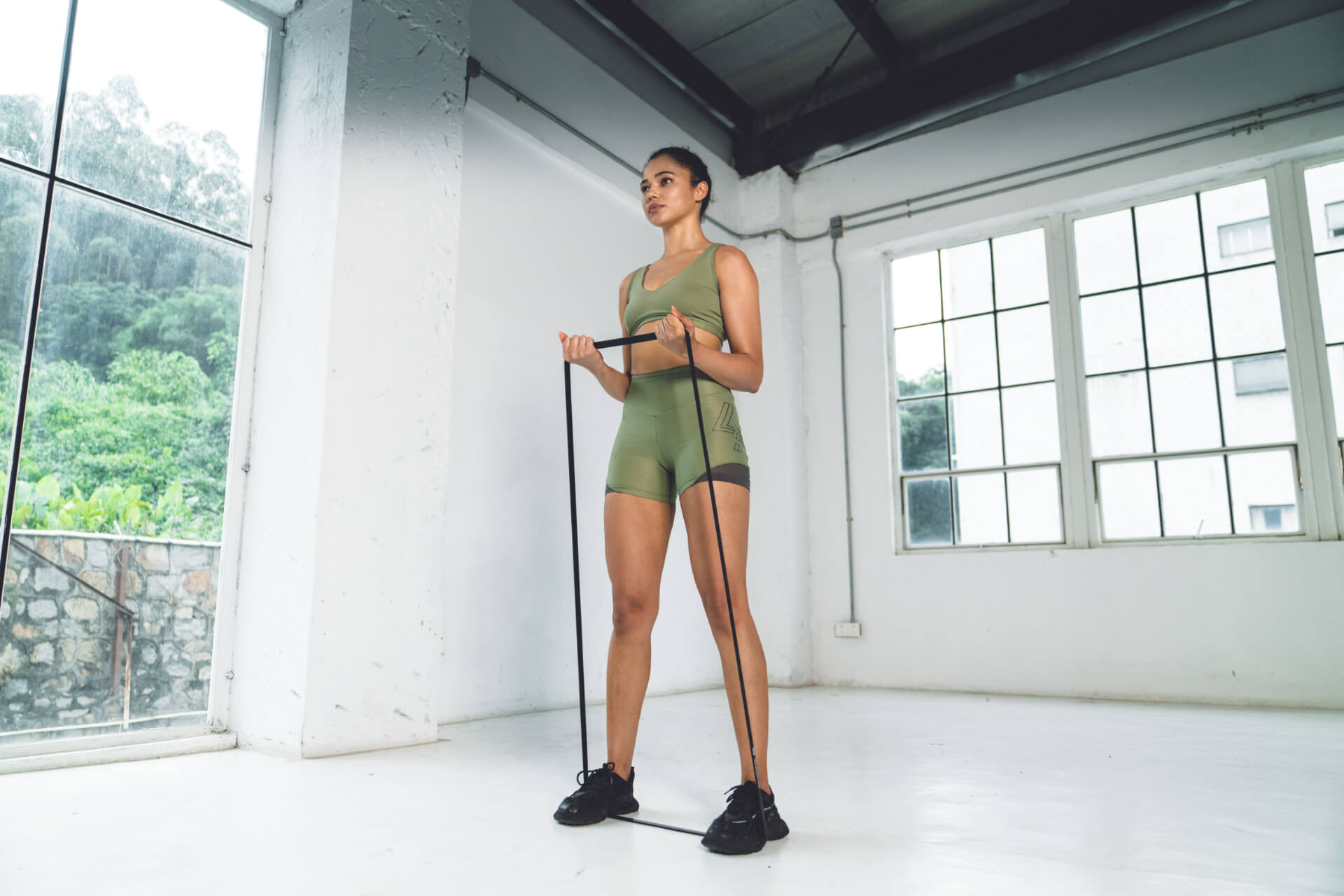 Young woman in olive green athletic wear using a resistance band for upper body exercises in a spacious, minimalist home gym